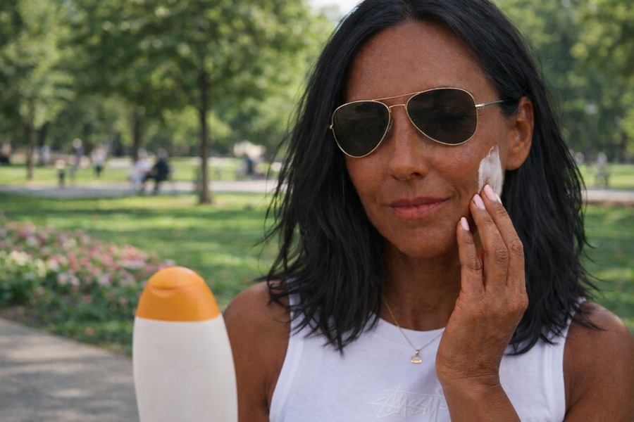 Mulher de óculos escuro e regata branca em área verde urbana passando protetor solar: cuidado pós carboxiterapia