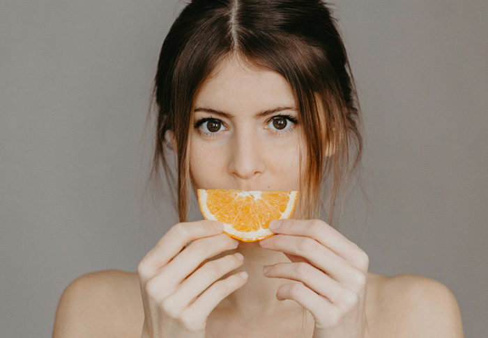 Mulher segurando uma fatia de laranja em frente a boca, olhando para frente.