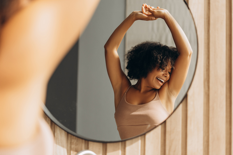 Mulher com autoestima se olhando no espelho, sorrindo, após sessão de massagem corporal.