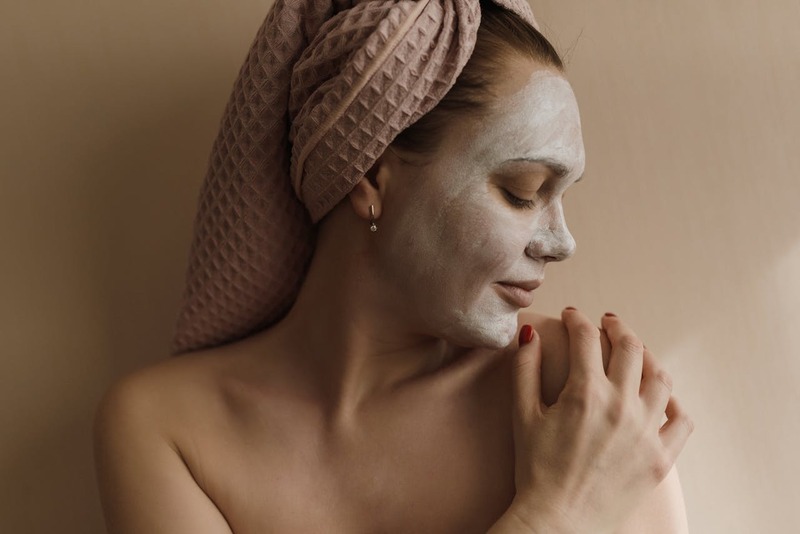 Mulher com toalha na cabeça e máscara facial hidratante aplicada no rosto, realizando cuidados com a pele, representando rotina de hidratação e skincare.