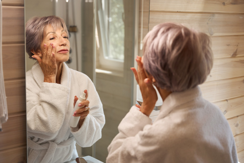Mulher madura realizando skincare em frente a espelho, para preservar a saúde da sua pele.