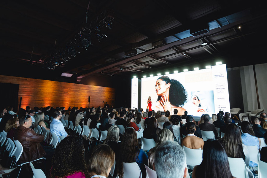 Imagem de auditório lotado, com apresentação sobre procedimentos estéticos sendo realizada por apresentadora no palco.
